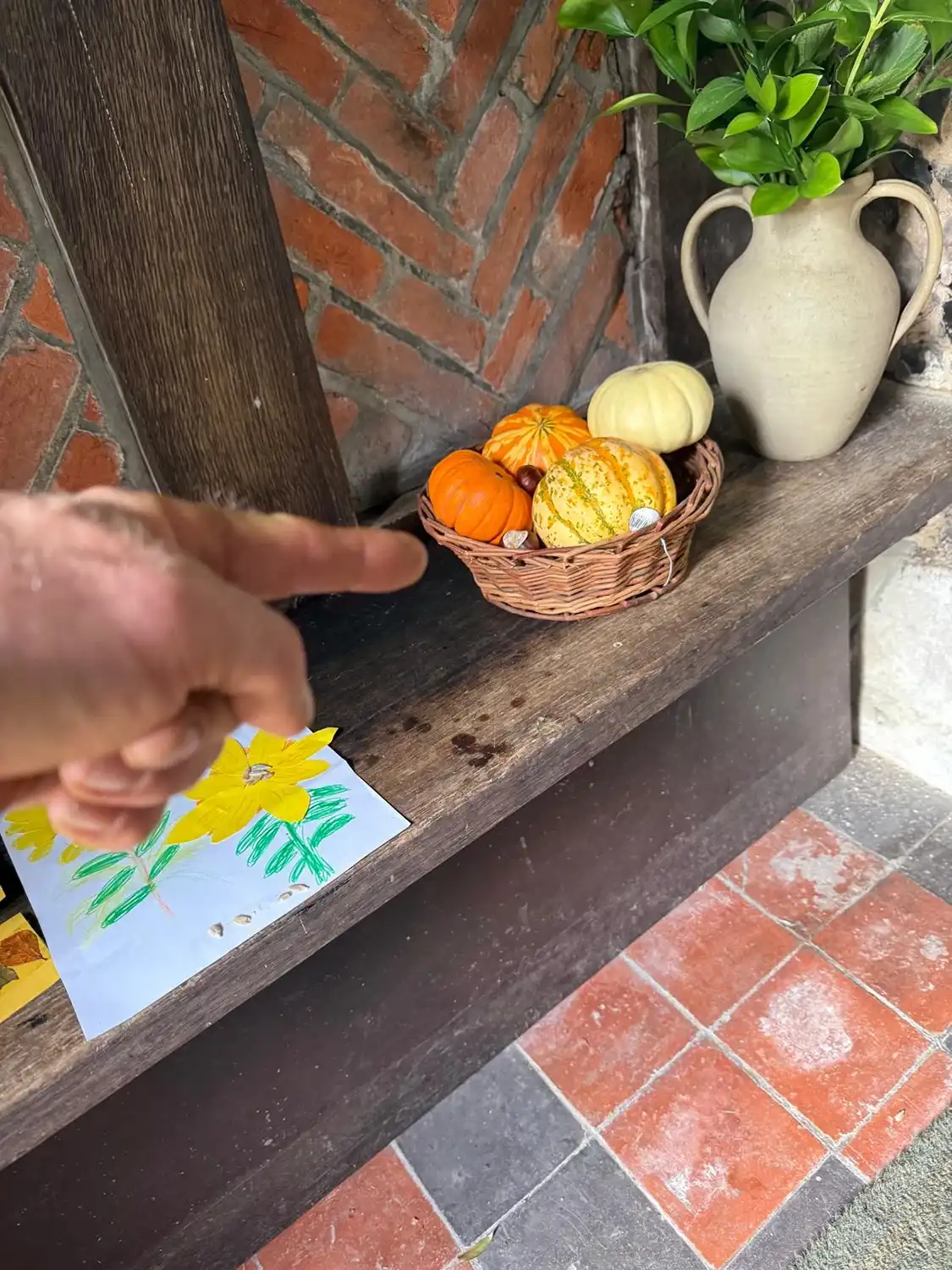 Hand pointing at a basket of small decorative pumpkins on a rustic wooden shelf with a vase and drawing.