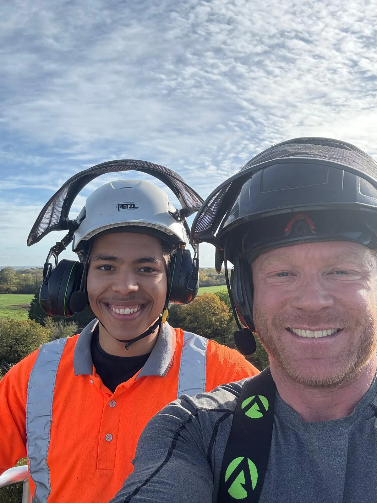 Two workers smiling wearing safety helmets and headsets with countryside background under a blue sky.