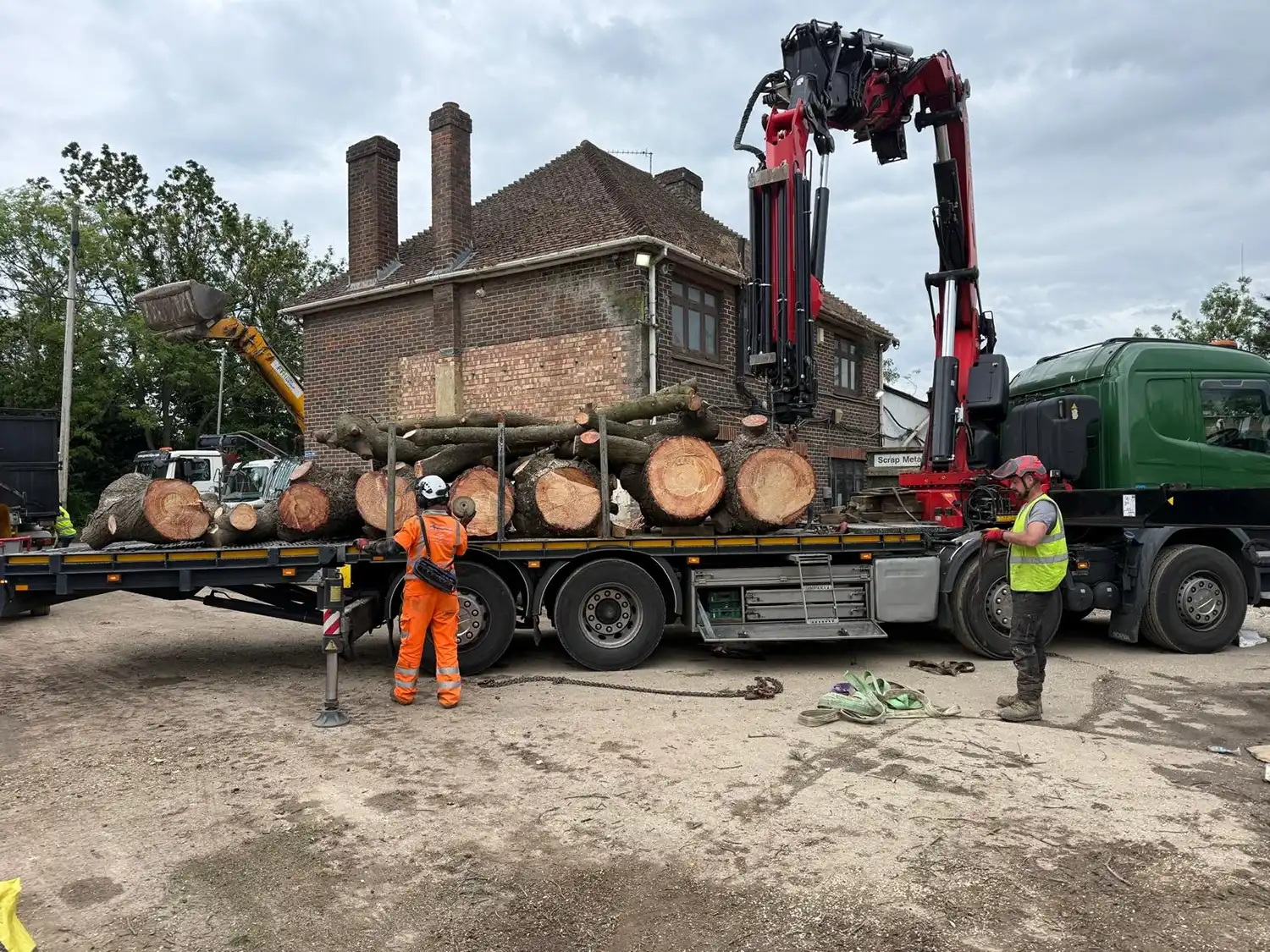 Two workers in helmets and safety gear load large logs onto a flatbed truck near a brick building.