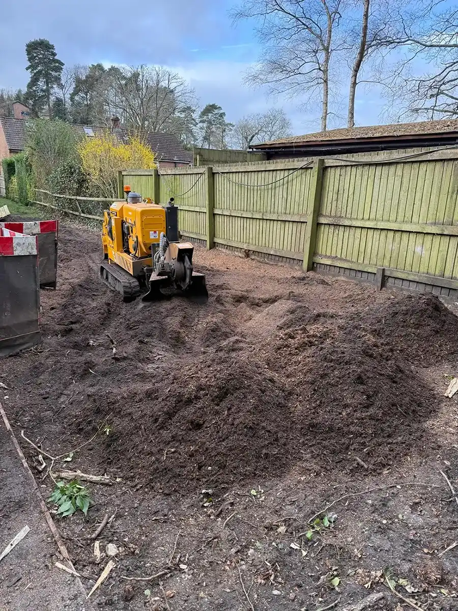 Small yellow digging machine working on dark soil near light wooden fence and trees.