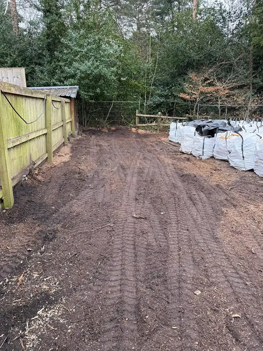 Dirt path with tire tracks between wooden fence on left and stacked white bags on right near trees.